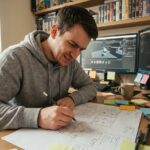 Jeune homme passionné esquissant le plan d'un niveau de jeu vidéo sur du papier millimétré dans un bureau maison avec des écrans de développement en arrière-plan